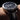 Black wristwatch with detailed face on a person's wrist against a dark background